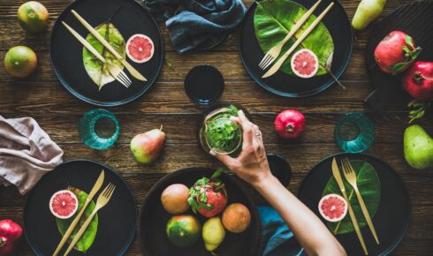 Autumn table styling for holiday dinner. Flat-lay of black dinnerware with fruit and leaves and female hands holding jug over rustic wooden table, top view. Preparing for Thanksgiving day or Christmas