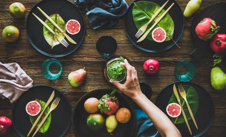 Autumn table styling for holiday dinner. Flat-lay of black dinnerware with fruit and leaves and female hands holding jug over rustic wooden table, top view. Preparing for Thanksgiving day or Christmas